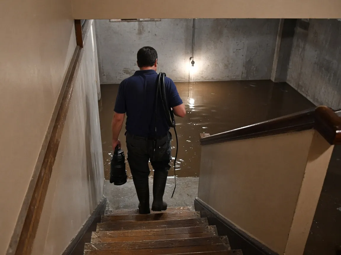 Technician carrying a submersible pump down the basement stairs toward standing water.